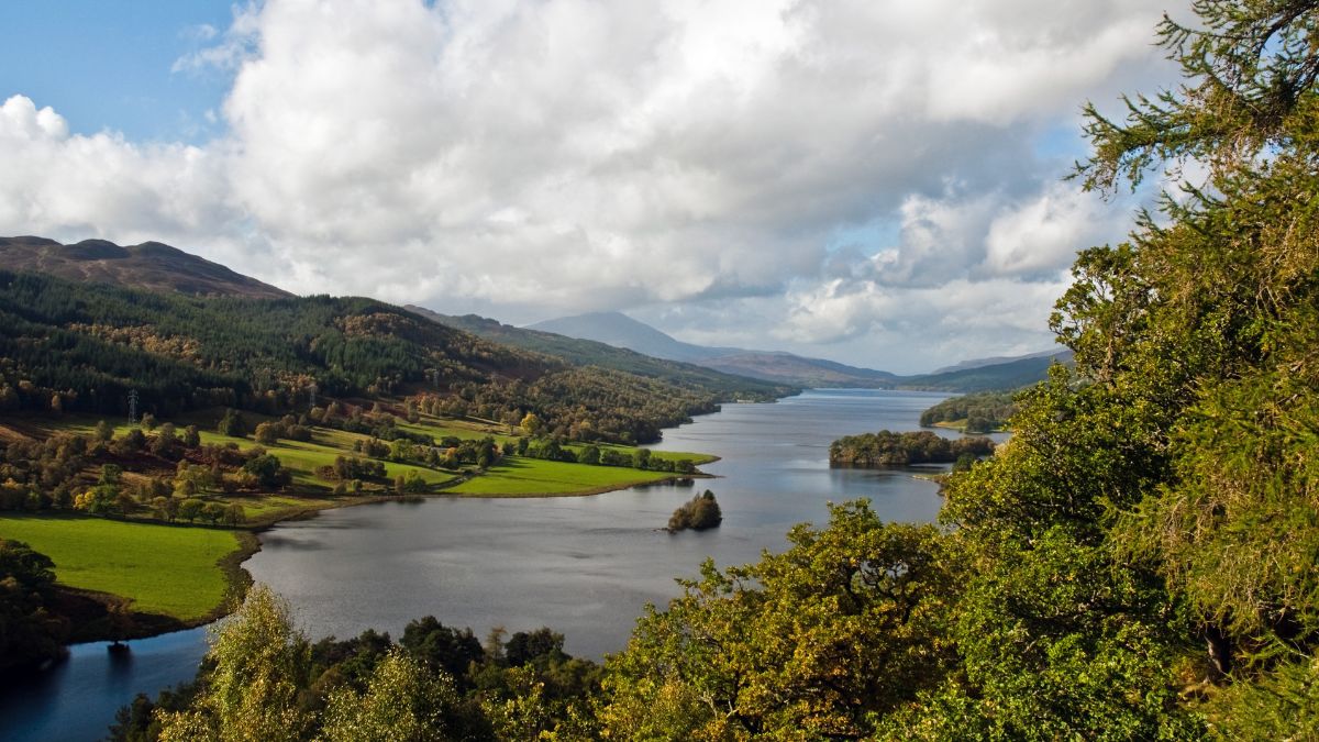 A scenic landscape featuring a calm lake surrounded by lush green fields and rolling hills, with distant mountains under a partly cloudy sky. Trees frame the view in the foreground.