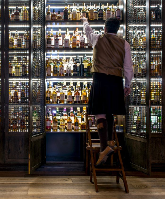 A man in traditional Scottish attire climbs a wooden step stool to reach a shelf filled with various bottles of whisky in a well-lit display cabinet.