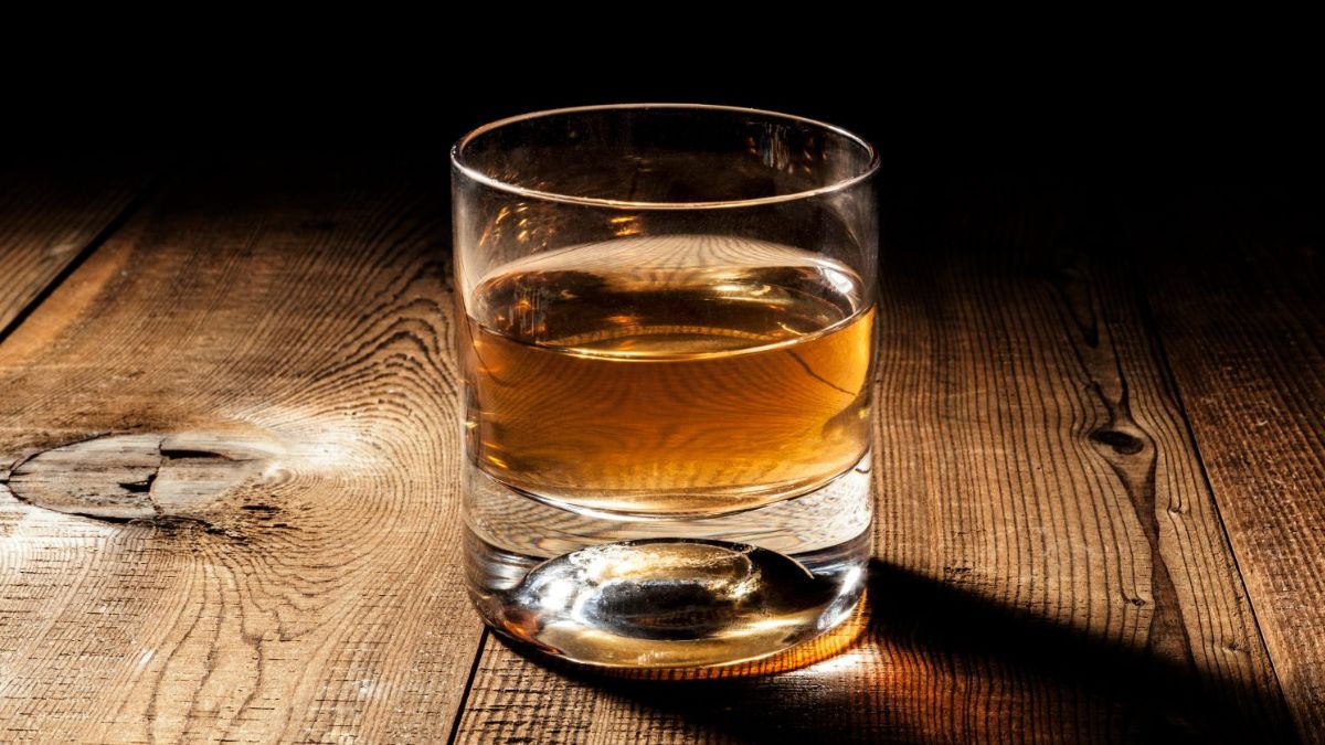 A glass filled with amber-colored whiskey sits on a rustic wooden surface, with dramatic lighting casting shadows.