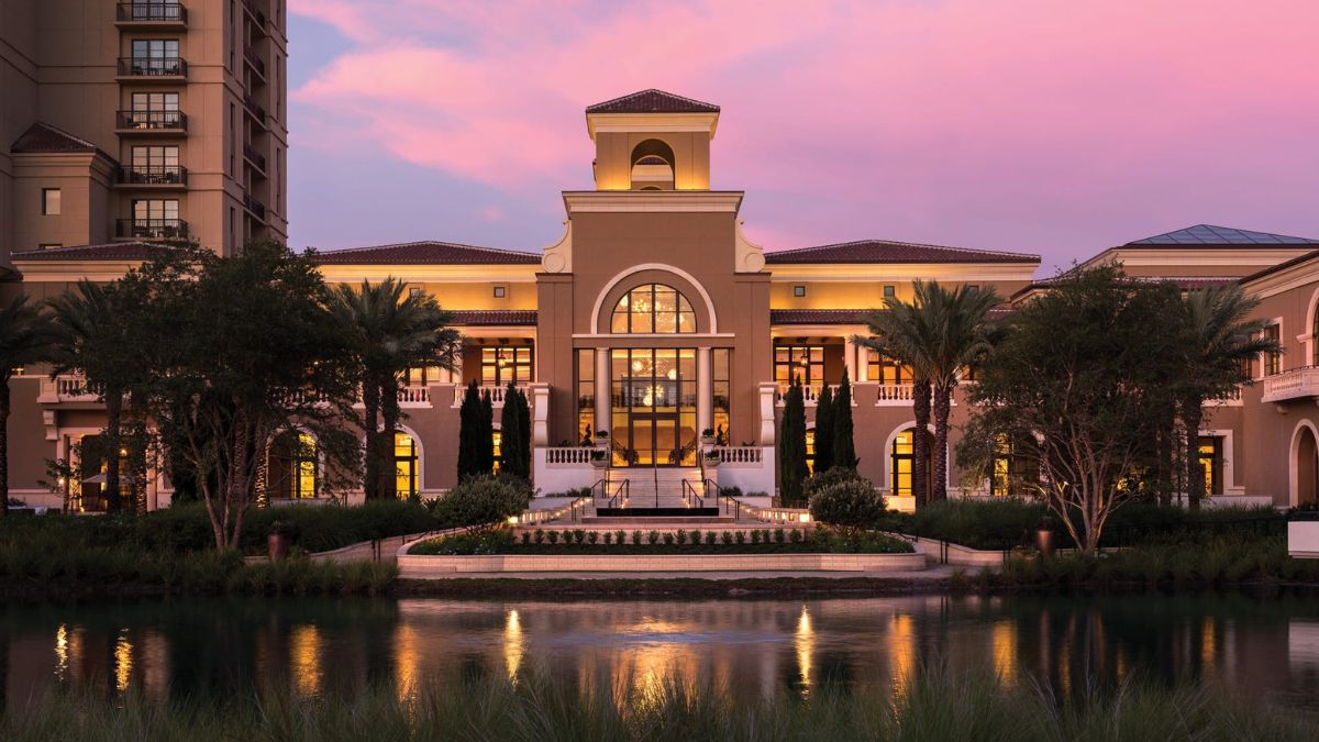 A luxurious hotel exterior at sunset, featuring elegant architecture, palm trees, and a tranquil water reflection. The sky is painted in soft pink hues.
