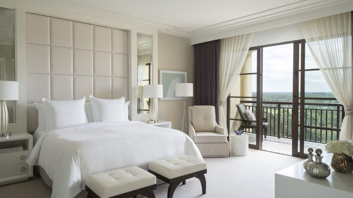 A luxurious hotel bedroom featuring a king-sized bed with white linens, two bedside tables with lamps, and a comfortable armchair. Large windows offer a view of the outdoors, with sheer curtains allowing natural light to fill the space. Elegant decor includes neutral tones and modern furnishings.