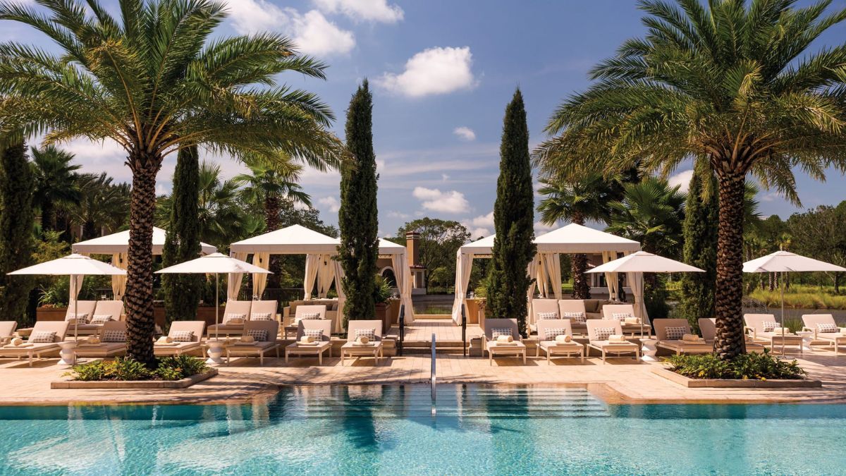 A luxurious pool area surrounded by palm trees and cabanas. Sun loungers line the poolside under large umbrellas, with a clear blue sky and a few fluffy clouds overhead.