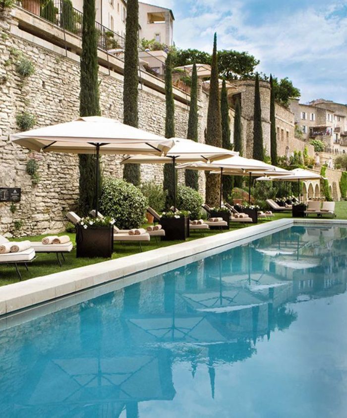 A serene poolside scene featuring lounge chairs under large umbrellas next to a crystal-clear pool. Tall cypress trees line a stone wall in the background, creating a tranquil atmosphere.