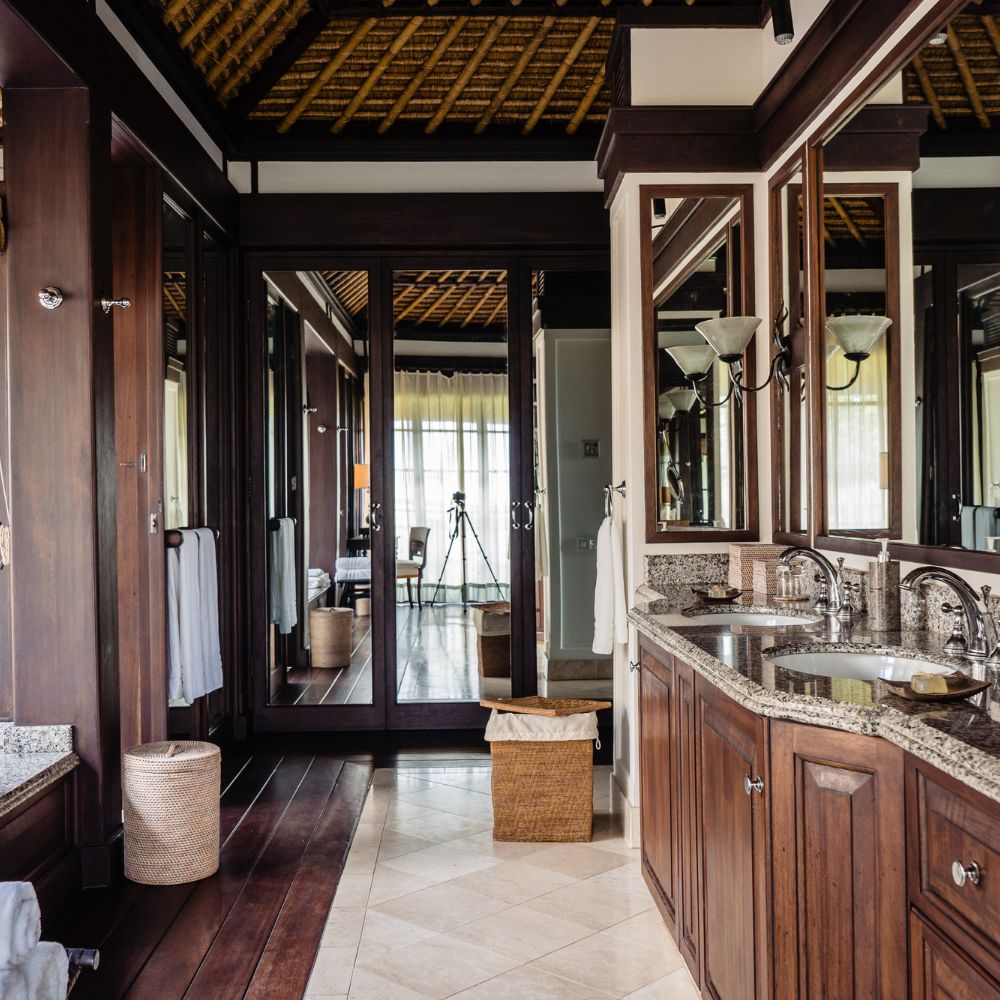 A spacious bathroom features dark wood cabinetry, a large mirror, and elegant light fixtures. Textured walls and a high, woven ceiling enhance the luxurious ambiance. A wicker laundry basket sits in the corner, and natural light floods the space from large windows.