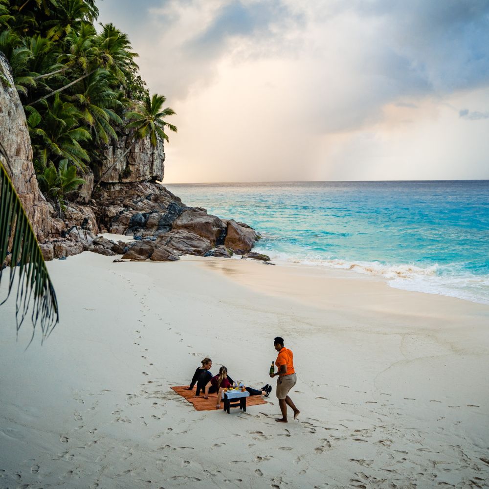 A serene beach scene featuring two people sitting on a wooden platform with a table, set against a backdrop of lush palm trees and rocky cliffs. A third person stands nearby, engaged in conversation. The ocean waves gently lap at the shore under a partly cloudy sky.