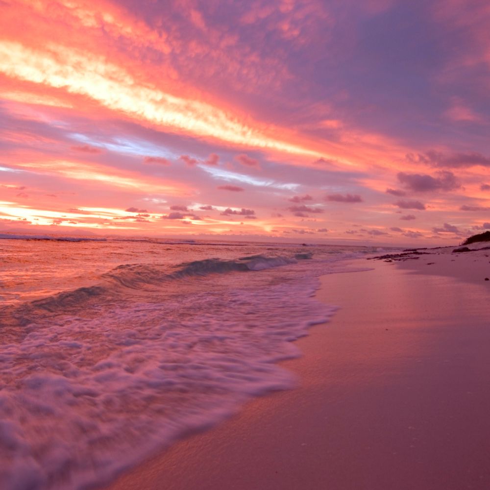A serene beach at sunset, with gentle waves lapping at the shore. The sky is painted in hues of pink, orange, and purple, reflecting beautifully on the water.