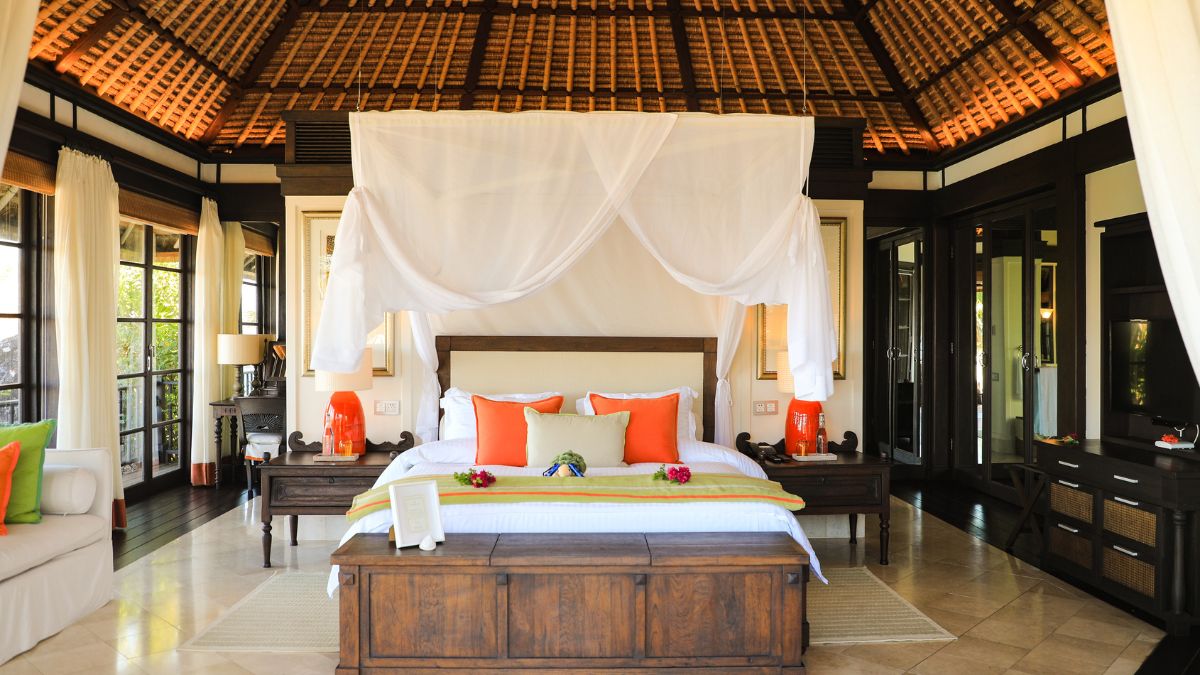 A spacious hotel room featuring a large wooden bed with white linens and bright orange pillows, draped with sheer white fabric. The room has tall ceilings with woven bamboo, large windows, and stylish decor including a cozy sitting area and decorative plants.