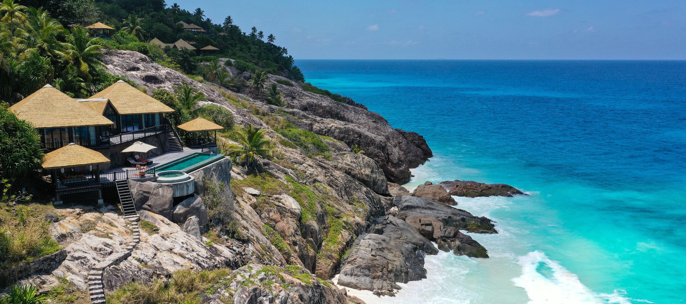 Luxurious villas perched on rocky cliffs overlooking a tranquil turquoise sea. Tropical palm trees surround the area, with a private infinity pool extending towards the ocean. Clear blue skies complement the serene landscape.
