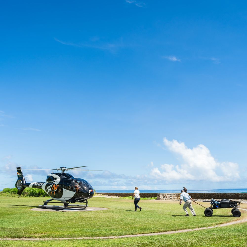 A helicopter is parked on a grassy area near the coast, with two people running towards it, one pulling a cart. The sky is bright blue with a few clouds.