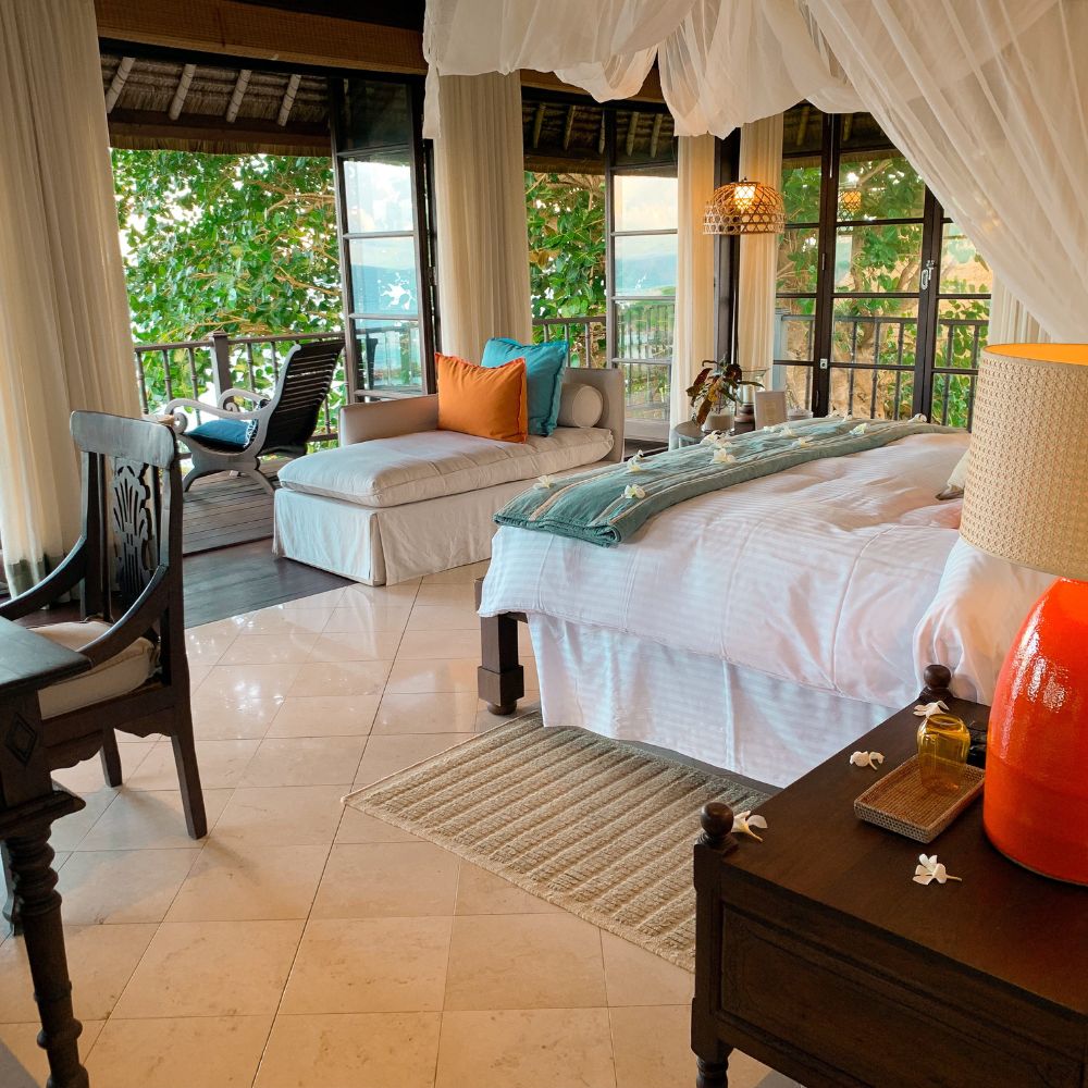 A cozy bedroom featuring a large bed with white linens, decorative pillows, and a turquoise throw. There’s a side table with a lamp, a wooden desk, and a lounge chair next to a window overlooking greenery. Sunlight filters through sheer curtains, creating a warm atmosphere.