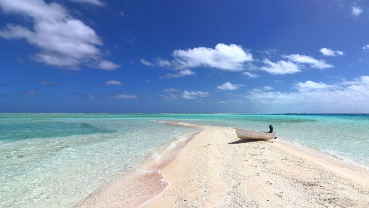 A small wooden boat rests on a sandy beach surrounded by clear turquoise waters under a bright blue sky with scattered clouds.
