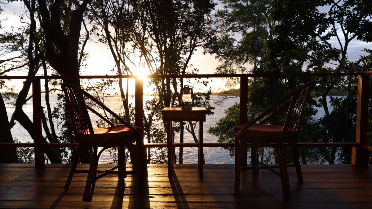 A serene balcony overlooking the water at sunset, featuring two wooden chairs and a small table, surrounded by lush trees. The sun casts a warm glow, creating a tranquil atmosphere.