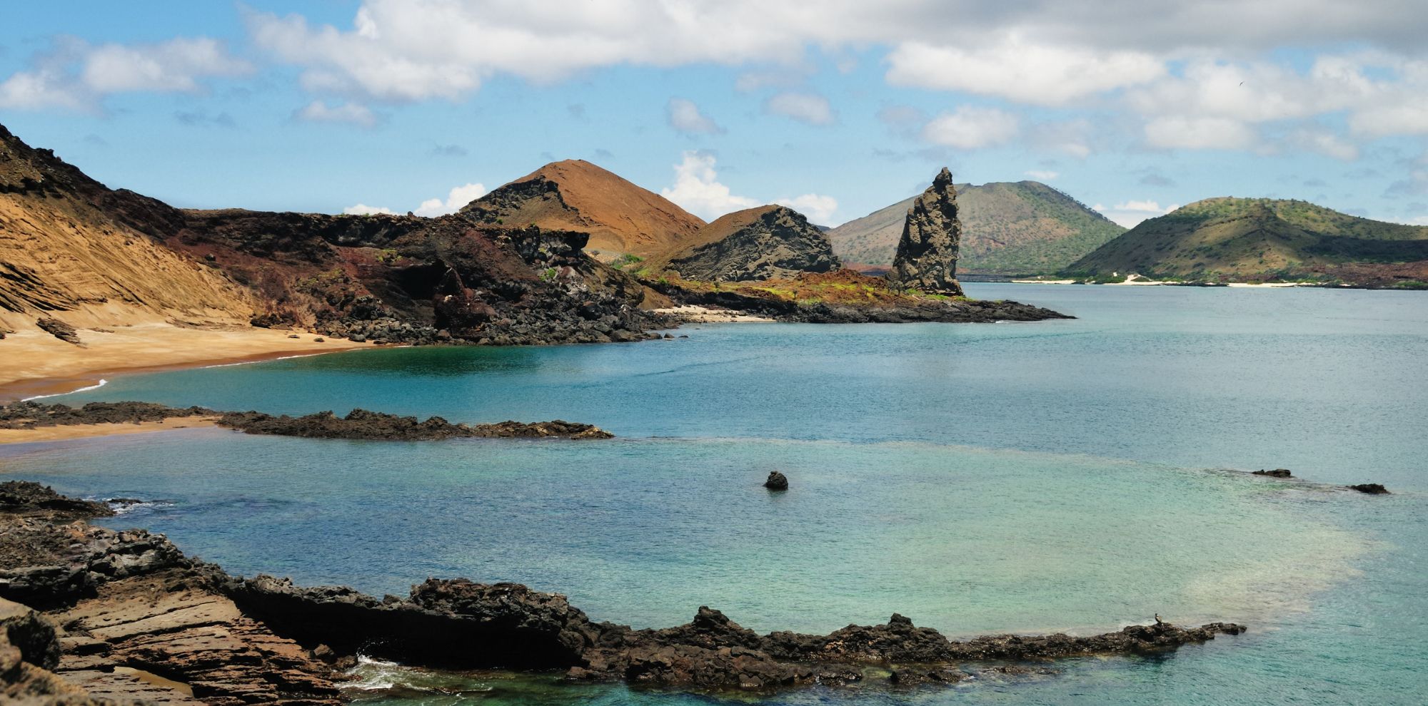 A serene coastal landscape featuring rocky shores, sandy beaches, and distant hills with varied vegetation. The water is clear, reflecting clouds and blue sky.