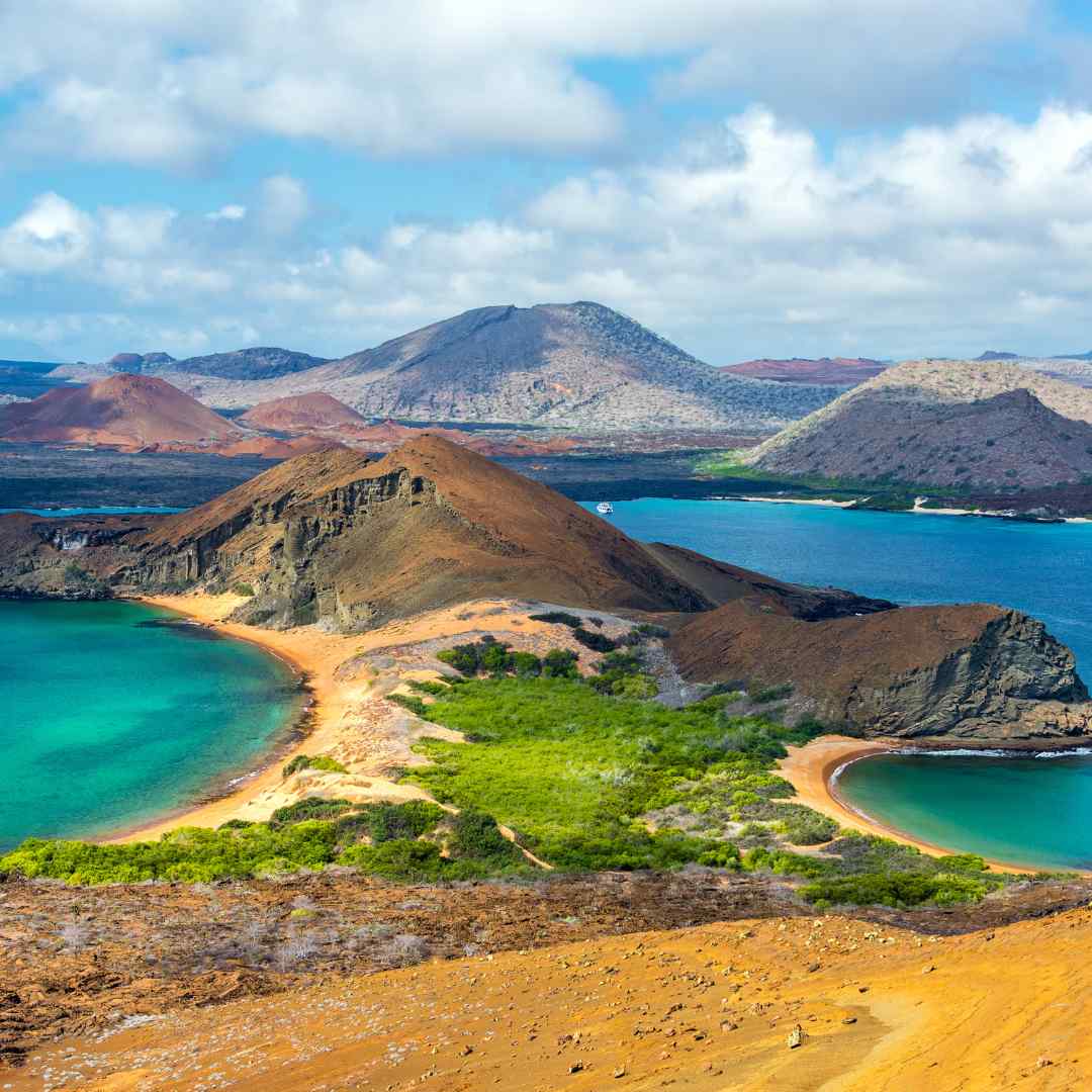 A stunning coastal landscape featuring a blend of turquoise waters, sandy beaches, and rugged volcanic hills. Lush greenery is visible along the shore, with distant mountains in the background under a partly cloudy sky.