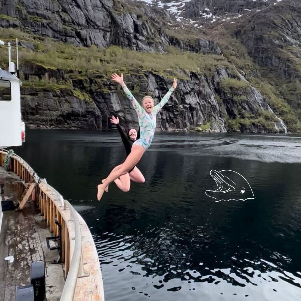 Two people jump joyfully off a boat into a calm, dark lake surrounded by steep, rocky cliffs and greenery. One person wears a light-colored swim suit while the other is in a black suit. A playful drawing of a fish is sketched nearby.
