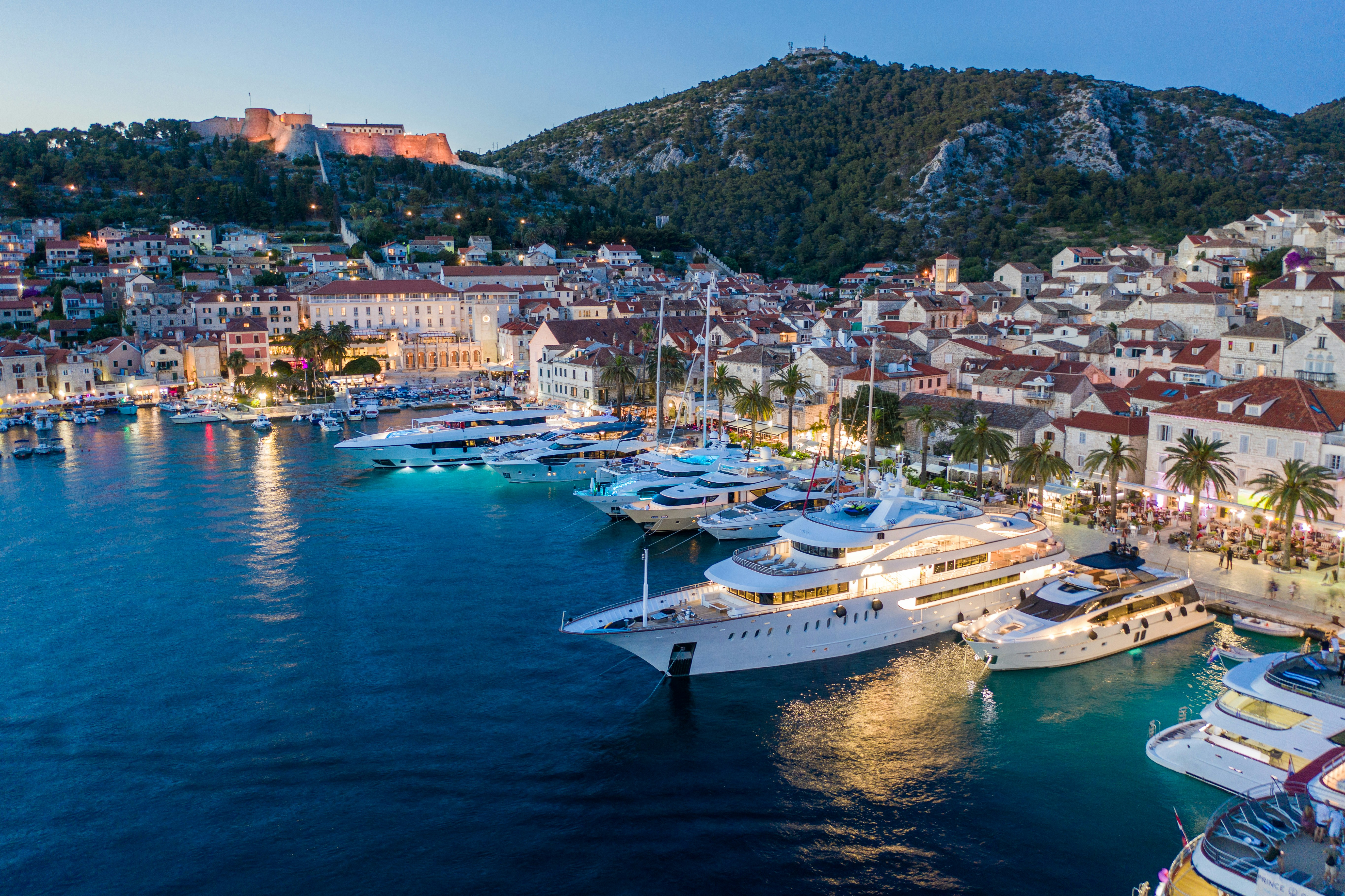 Aerial view of a Mediterranean harbor at twilight, with luxury yachts docked along a stone waterfront lined with buildings and palm trees.
