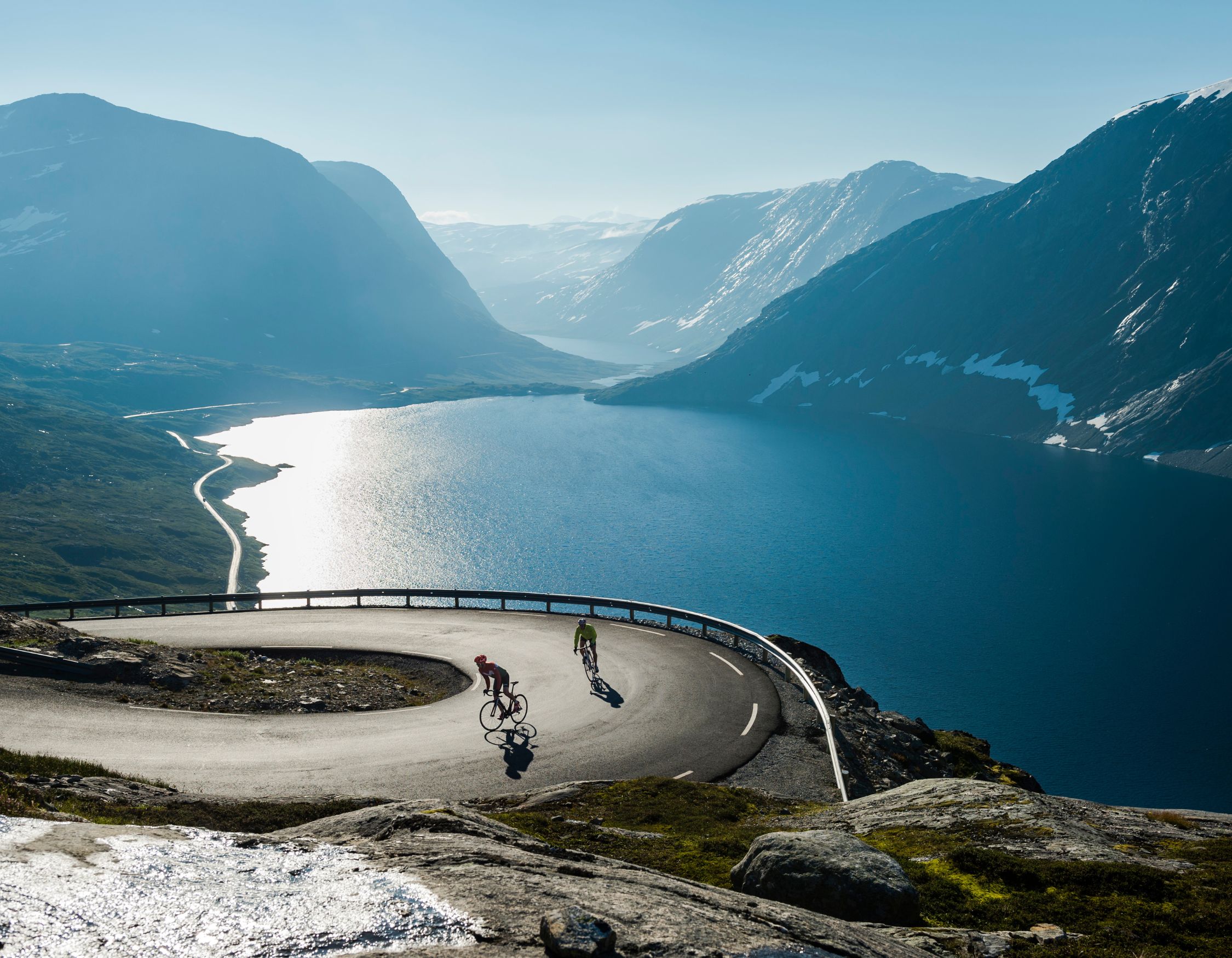 Two cyclists navigate a winding road along a scenic mountain landscape, with majestic peaks and a serene lake reflecting the blue sky in the background.