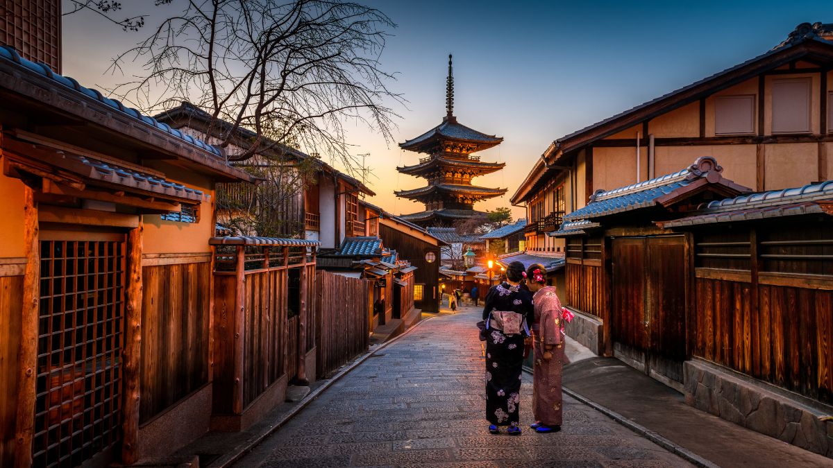 Two women in traditional kimonos stand in a narrow, cobblestone street at sunset. The backdrop features a pagoda and wooden buildings, with warm orange and blue hues in the sky.