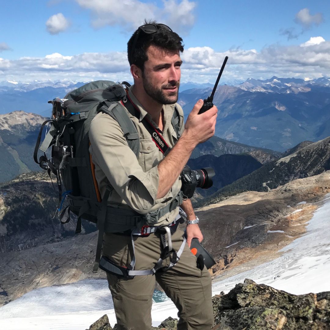 A man with a backpack and camera on his chest stands on a rocky mountain peak, holding a walkie-talkie. He is dressed in outdoor gear and surrounded by a scenic landscape of mountains and a glacier under a cloudy sky.