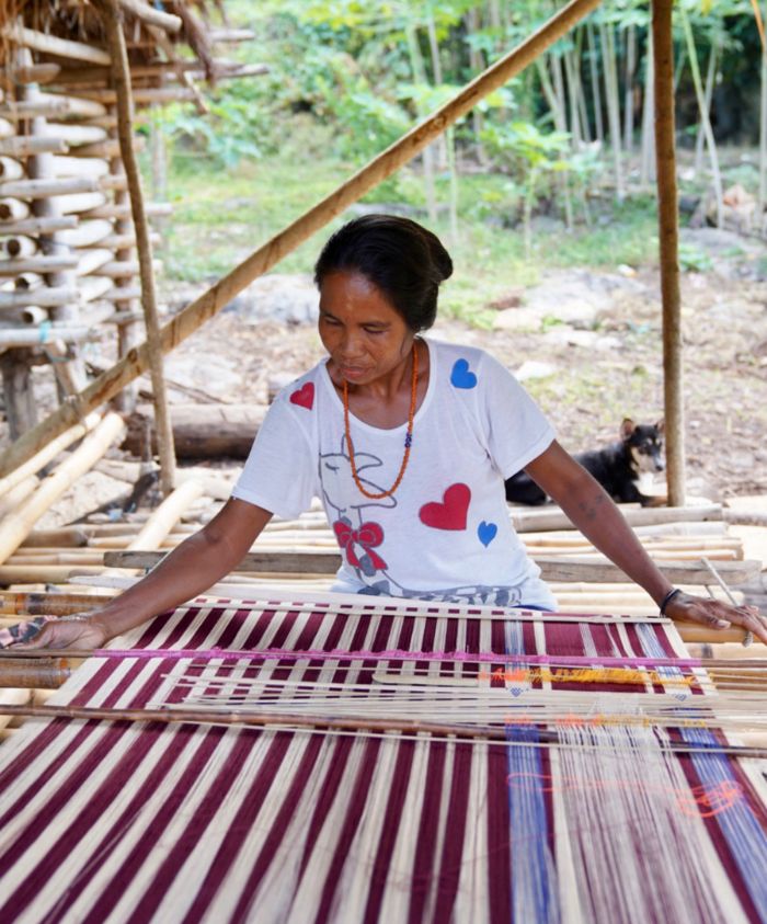 A woman weaves fabric on a traditional loom in a rural setting. She wears a white t-shirt with red and blue heart patterns and is focused on her craft, surrounded by bamboo structures and greenery. A dog is visible in the background.
