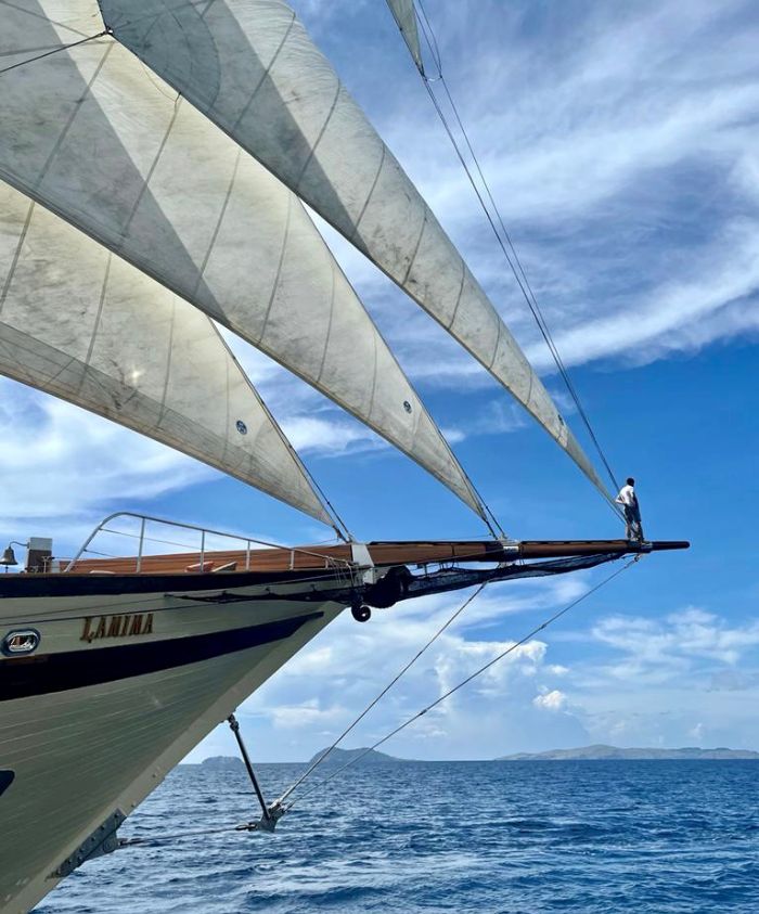 A sailboat with large white sails is cutting through the blue ocean. A person stands at the bow, looking out at the horizon under a partly cloudy sky.