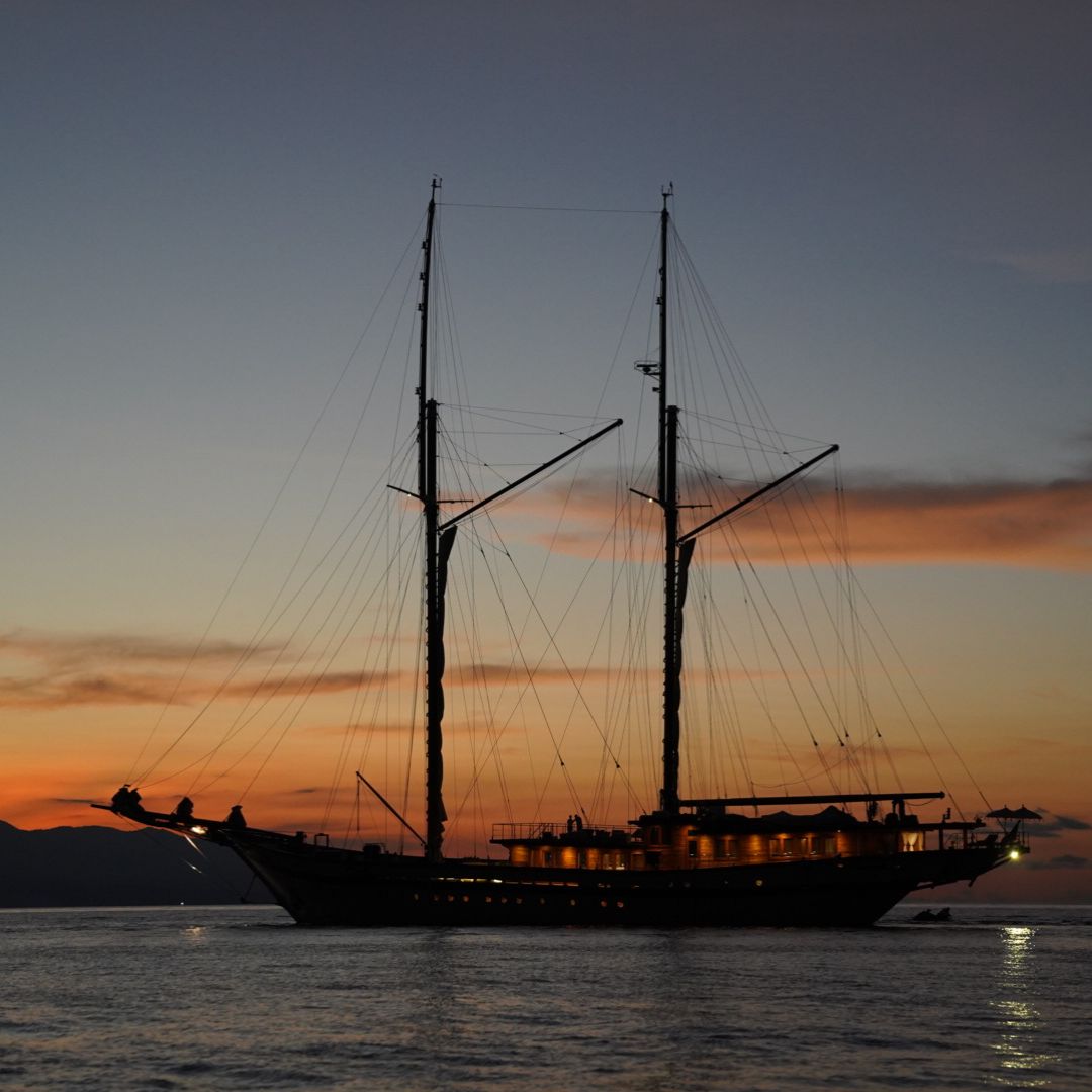 A majestic sailing ship with tall masts silhouetted against a colorful sunset sky. The vessel is illuminated from within, casting a warm glow on the calm waters below.