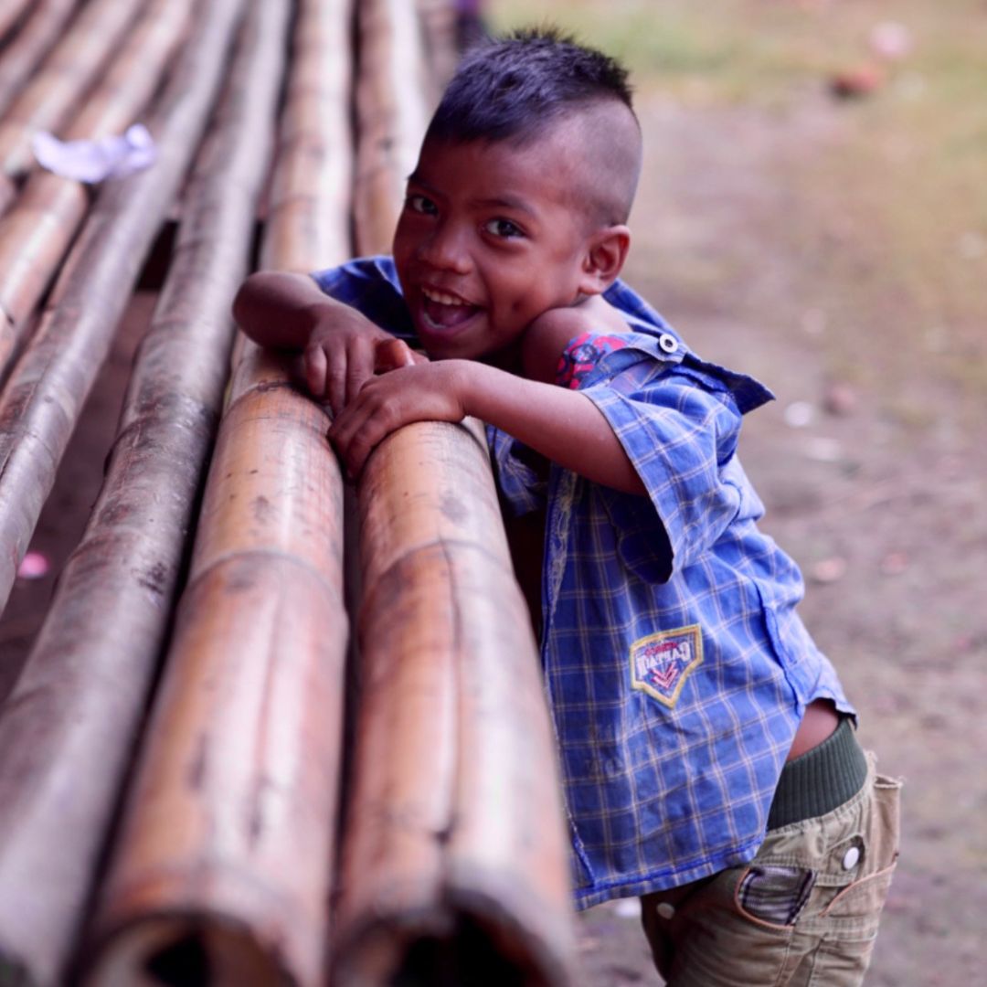 A young boy with a mohawk hairstyle smiles while playfully leaning against a stack of bamboo. He is wearing a blue checked shirt and green pants. The background features a blurred outdoor setting.