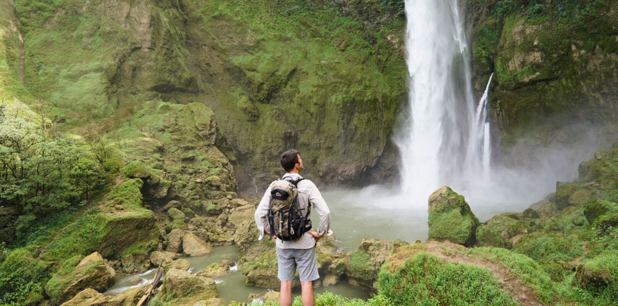 A person stands on rocky terrain, gazing at a large waterfall cascading down a lush, green cliff. The scene is surrounded by dense vegetation and boulders.