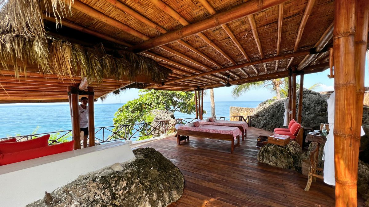 A spacious outdoor seating area with bamboo and thatched roofing overlooking the ocean. The wooden floor features a pink bed, lounge chairs, and decorative rocks, surrounded by tropical greenery.