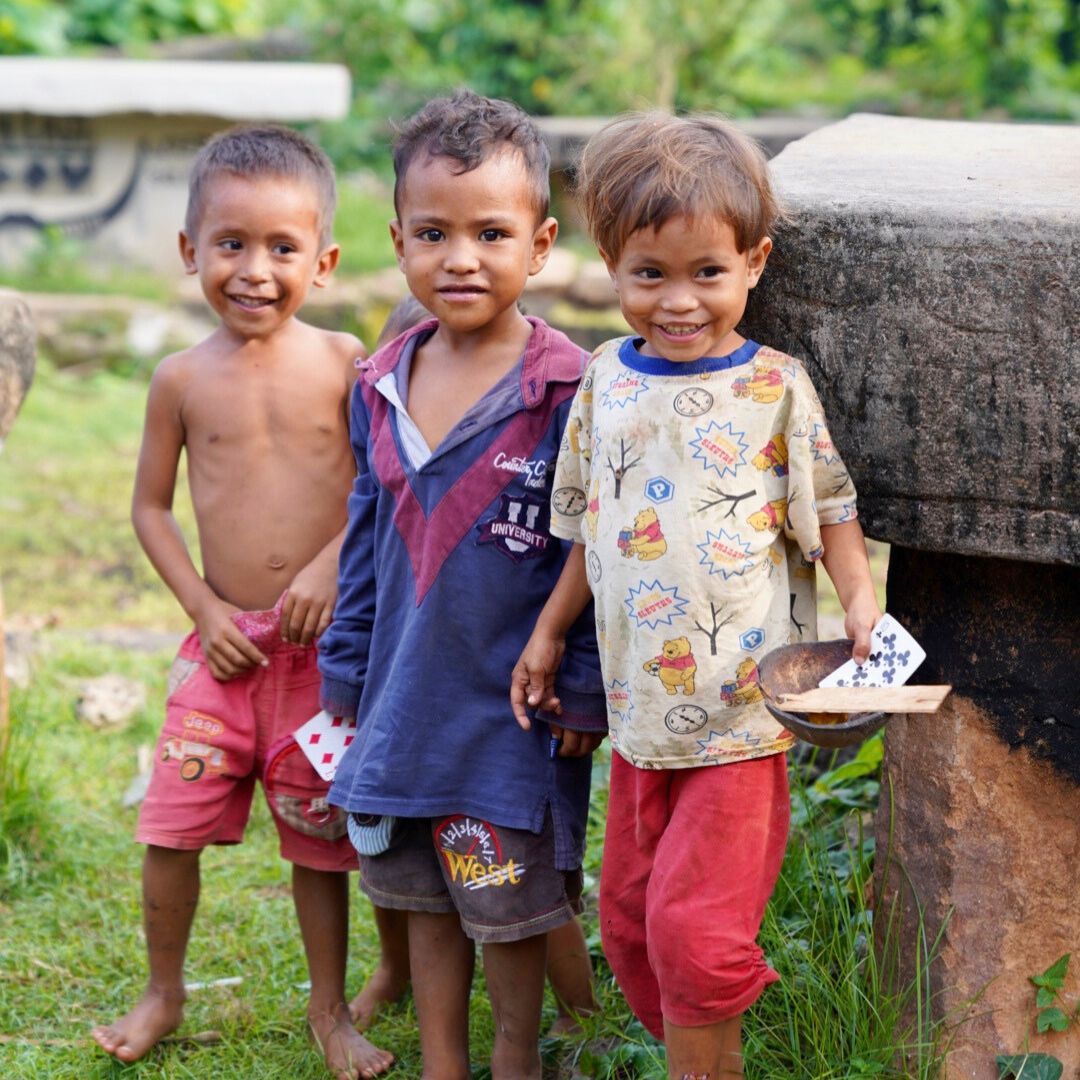 Three smiling young boys standing outdoors, dressed in colorful, worn clothes. They are positioned playfully next to a stone structure, with greenery in the background.