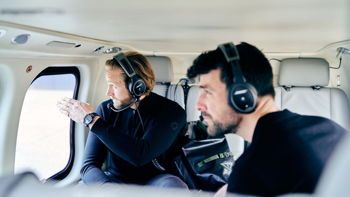 Two men wearing headsets sit inside a helicopter, looking out of the window. One points outside, while the other focuses intently. The interior is modern and streamlined, with minimalistic seating.