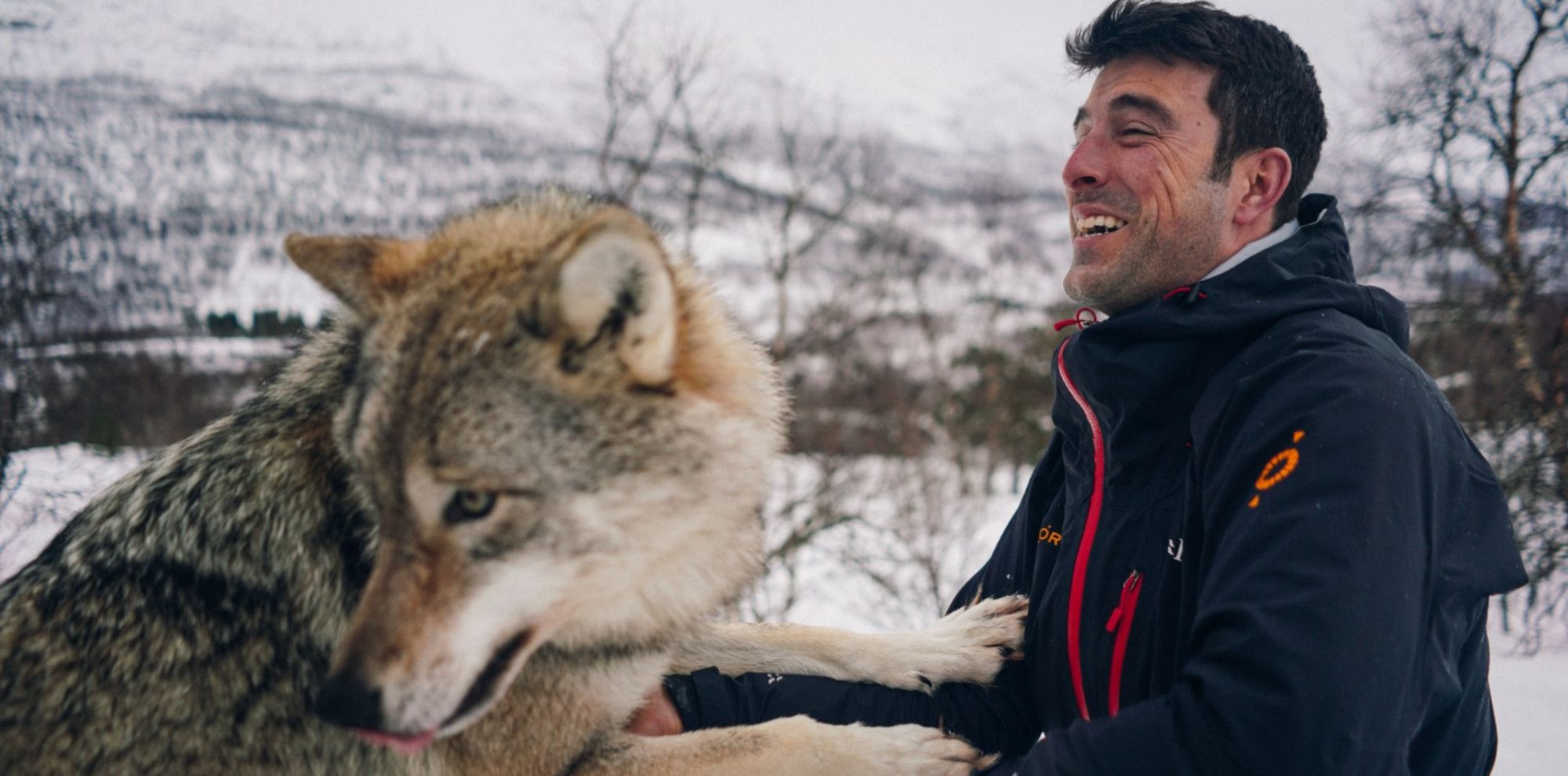 A man in winter clothing smiles joyfully as he interacts closely with a large wolf in a snowy landscape. Trees and distant mountains are visible in the background.