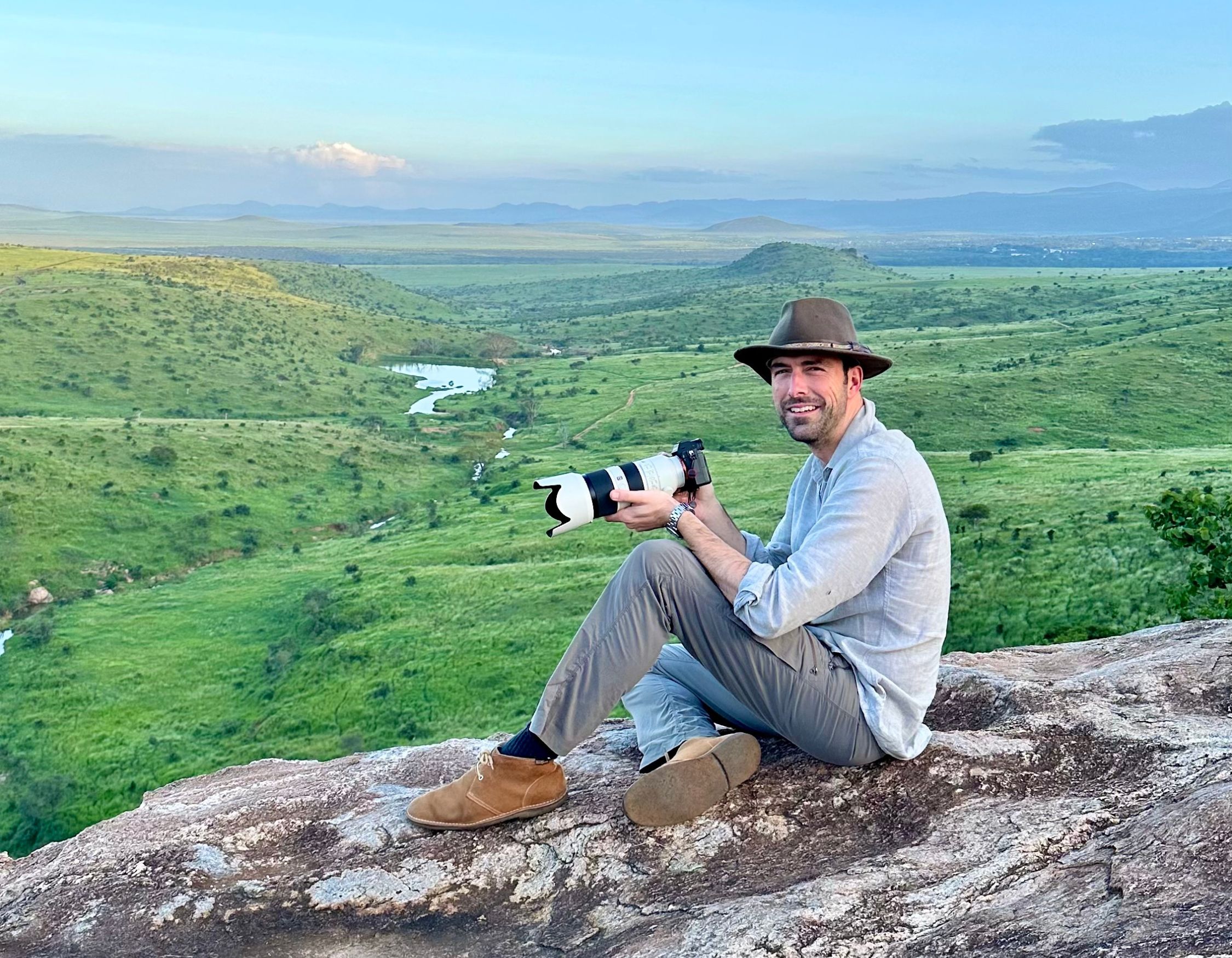 A Pelorus expert travel designer in a hat sitting on a rock holding a professional camera with a long lens, overlooking a lush green valley.