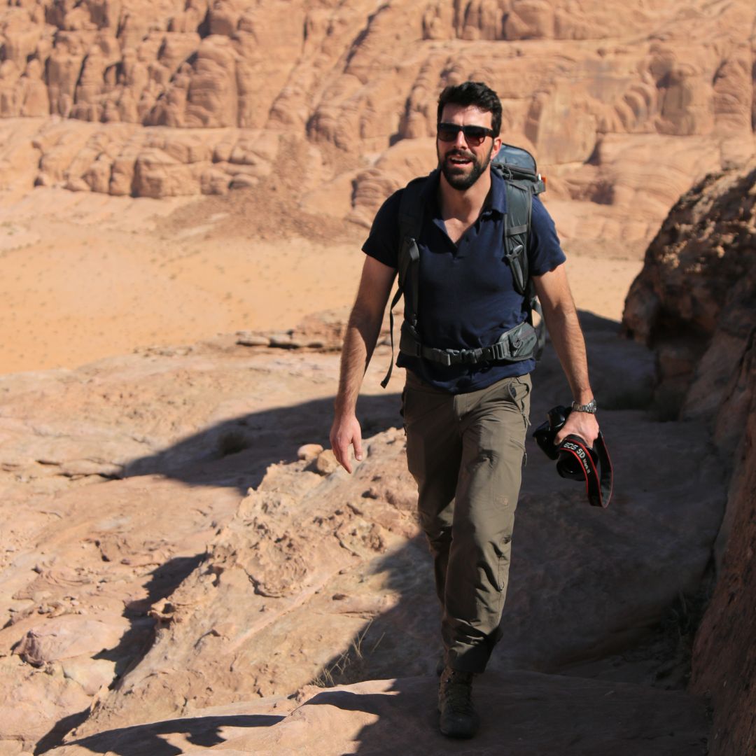 A man hiking through a rocky desert landscape, wearing a backpack and holding a camera. He has a beard and sunglasses, with sunlit rock formations in the background.