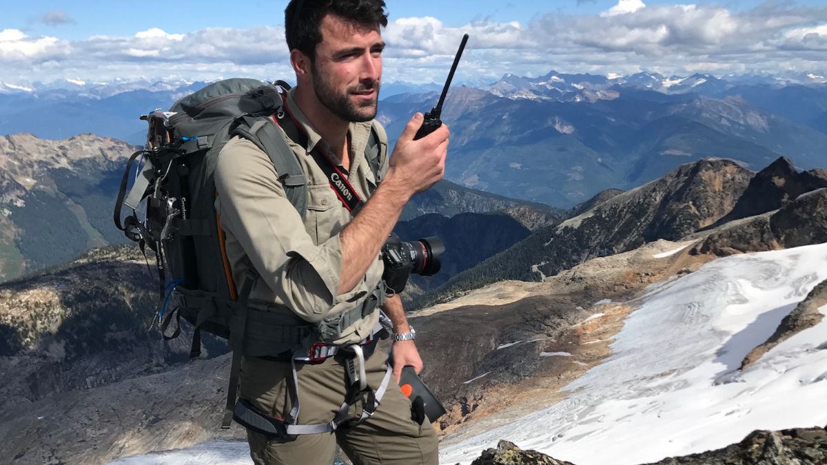 A man with a backpack and camera stands on a mountain summit, using a walkie-talkie while surrounded by a panoramic view of mountains and valleys.