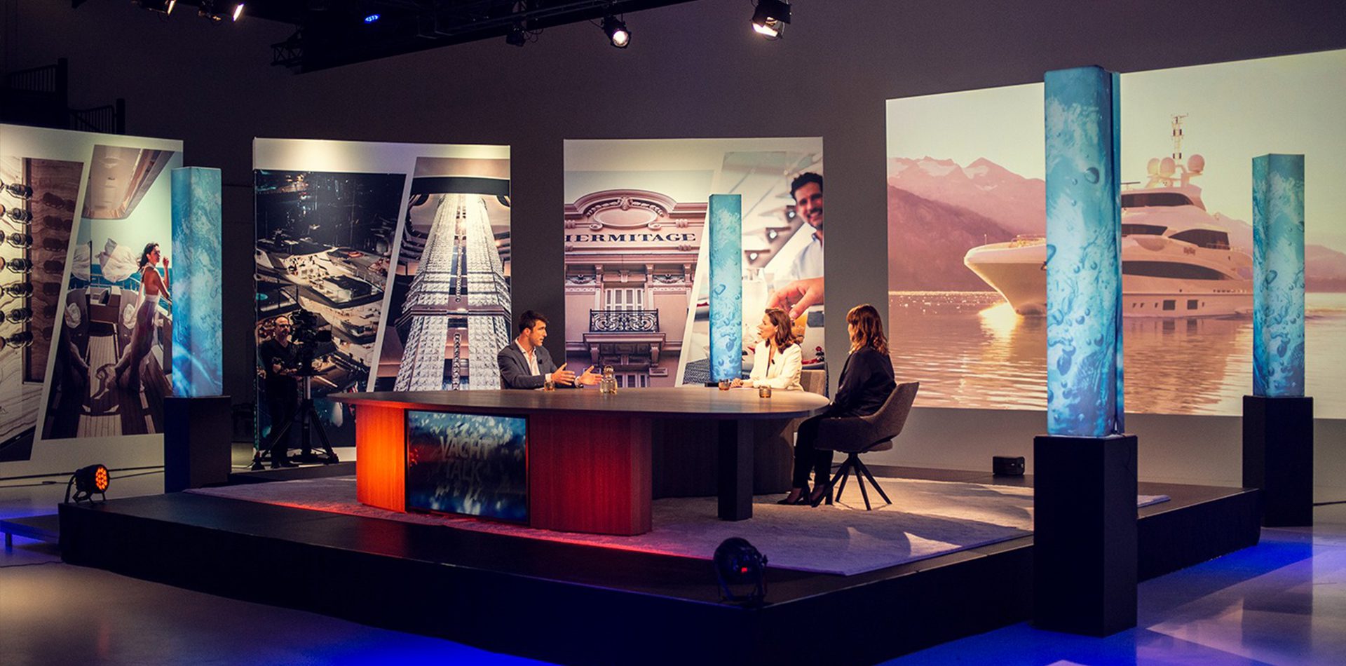 A television studio set featuring a central discussion table with three people seated. Behind them are large backdrop images of luxury yachts and scenic mountains, with illuminated blue columns flanking the sides. Lighting creates a modern, professional atmosphere.