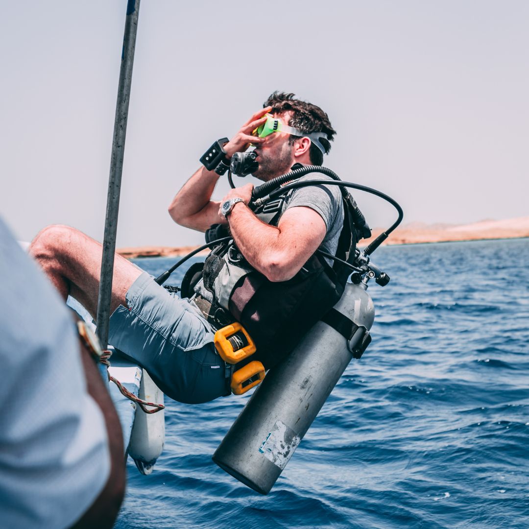 A man wearing a diving mask and snorkel is preparing to dive into the water, holding onto a boat's ladder. He has a scuba tank on his back and appears focused and ready for underwater exploration. The clear blue water surrounds him, with a hint of land in the background.