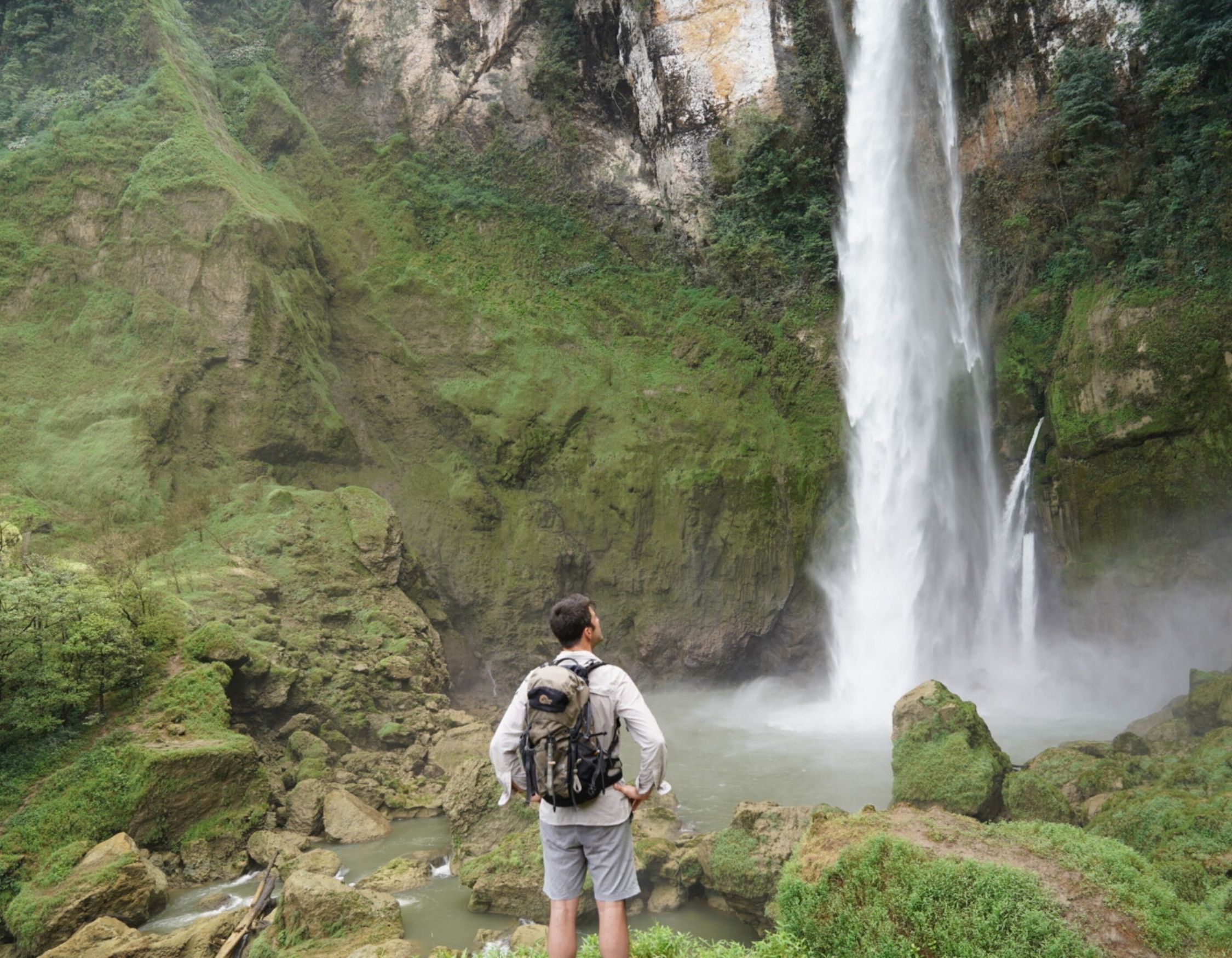A hiker with a backpack stands on a rocky ledge, looking up at a massive, powerful waterfall cascading down a green cliff.