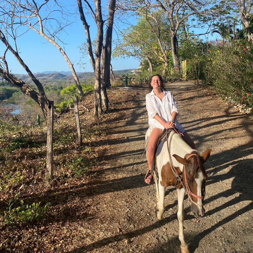 A person riding a brown and white cow along a scenic dirt path surrounded by trees and greenery, smiling up at the sky.