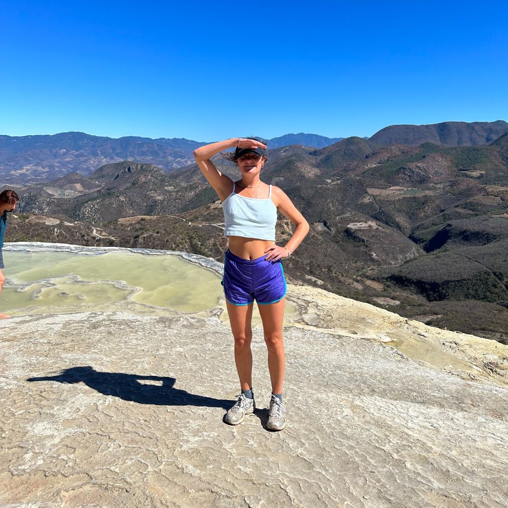 A woman stands on a rocky surface with her hand shielding her eyes from the sun, overlooking a mountainous landscape. She is wearing a light blue crop top and purple shorts. In the background, blue skies and green hills create a scenic view.