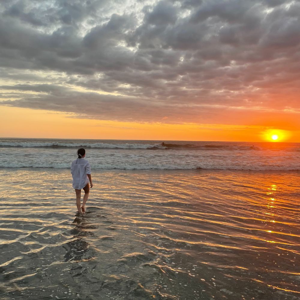 A person walks barefoot along a wet beach at sunset, with waves gently lapping at their feet and vibrant orange and yellow hues filling the sky.