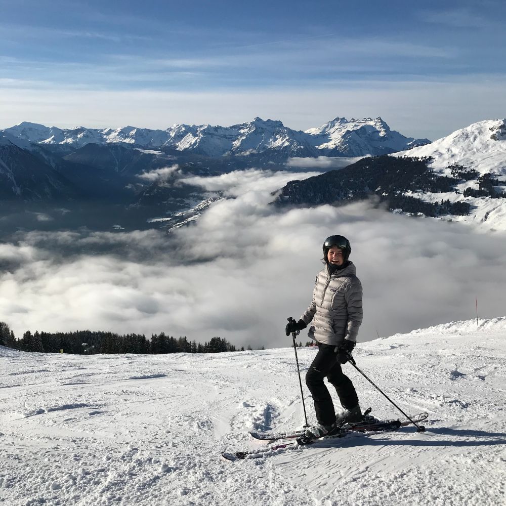 A skier in winter gear poses on a snowy slope with majestic mountains and a sea of clouds in the background.