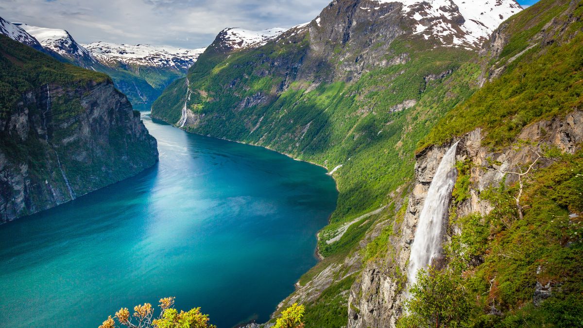 A stunning view of a deep blue fjord surrounded by steep, green cliffs and majestic snow-capped mountains. A waterfall cascades down the rocky cliff into the fjord below.