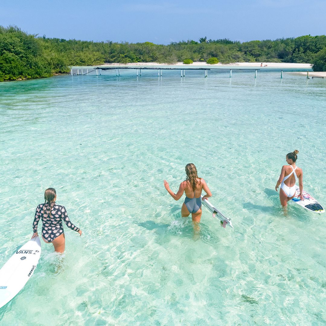Three women wade through clear turquoise water towards a sandy shore, carrying surfboards. Lush greenery and a wooden walkway are visible in the background. The sky is bright and sunny.