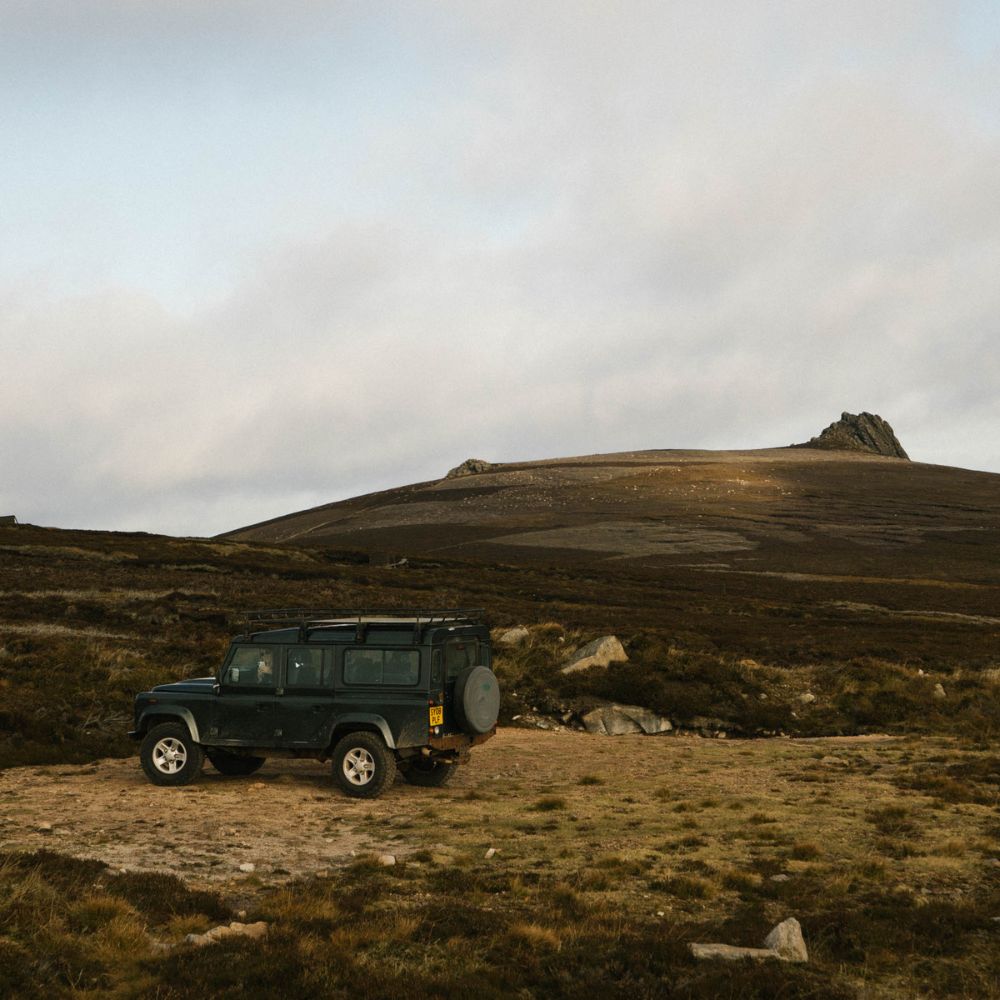 A green SUV parked on a dirt path in a rugged landscape, with rolling hills in the background and a rocky outcrop illuminated by soft sunlight.