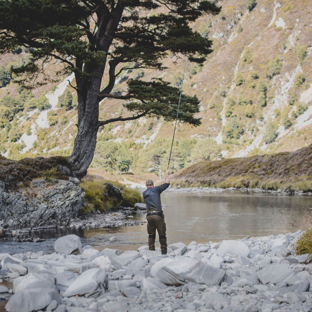 A fisherman stands in a river, casting his line amid a serene landscape of rocky shores and lush, green hills in the background. A tall tree provides shade along the water's edge.
