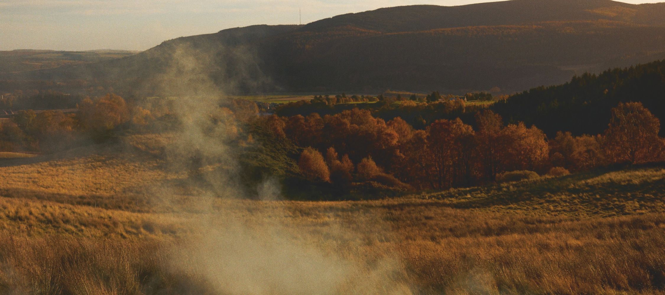 A serene landscape features rolling hills and a valley, bathed in warm autumn colours. Trees with orange and golden leaves dot the scene, while mist rises softly from the ground, creating a mystical atmosphere.
