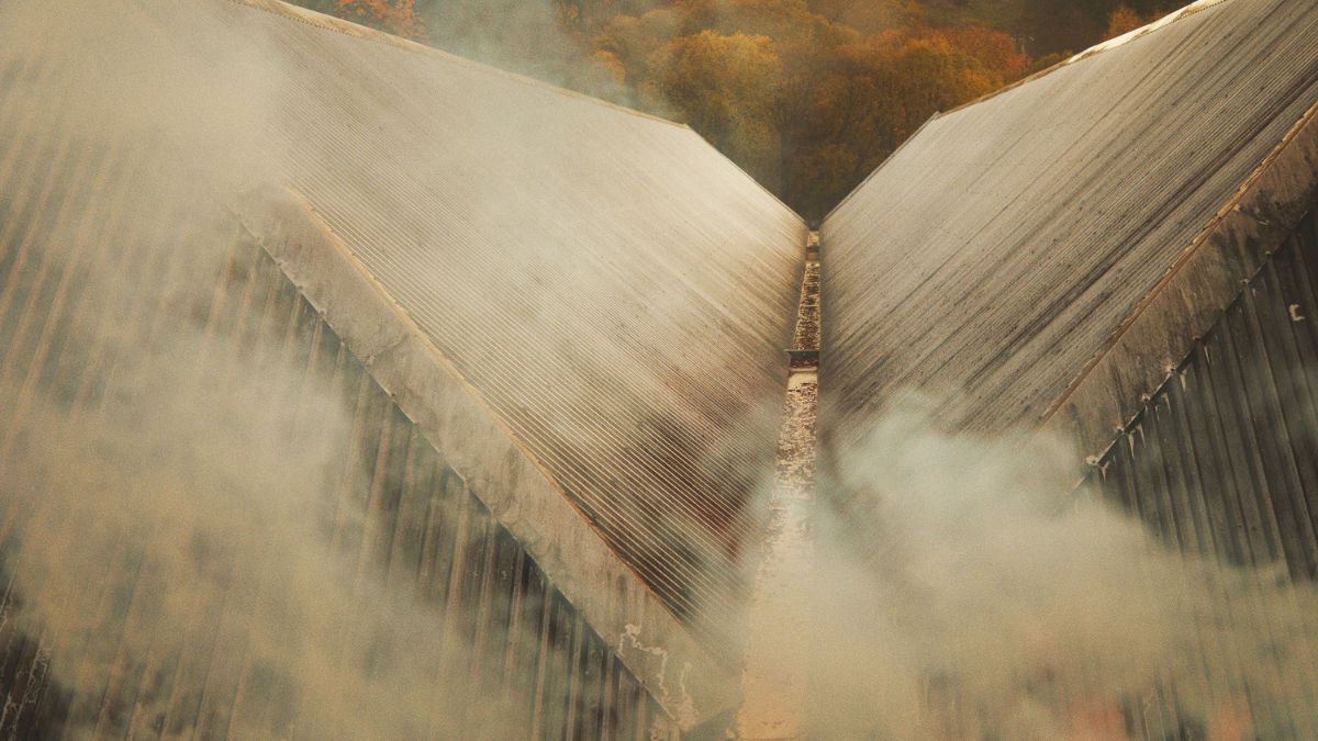 Aerial view of two triangular rooftops meeting at a central point, surrounded by wisps of smoke and overlooking a forested area with autumn foliage.