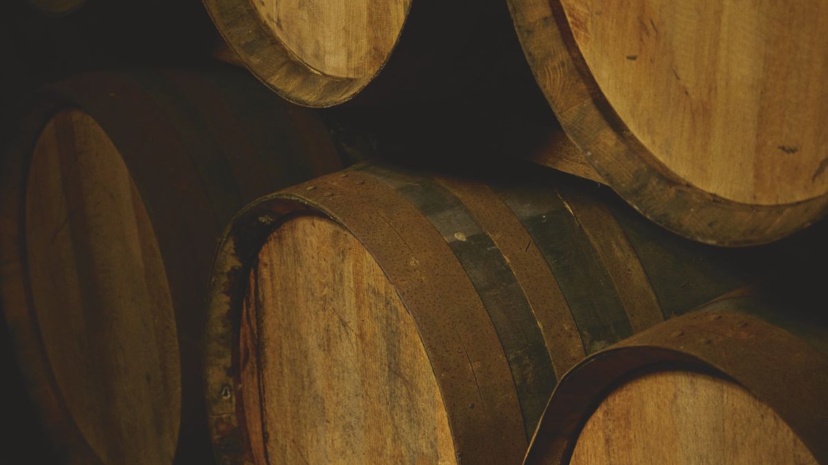 Close-up of several wooden wine barrels stacked against each other in a dimly lit cellar. The barrels have dark metal bands and a rustic finish, showcasing the natural wood grain.