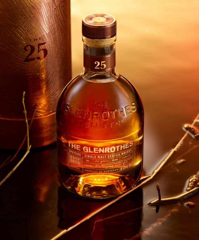 A bottle of The Glenrothes 25 Year Old single malt Scotch whisky, featuring a round shape and a cork stopper, sits on a reflective surface. The background displays warm golden tones, enhancing the rich amber color of the whisky. A partially visible elegant box accompanies the bottle, and some natural elements like twigs are scattered nearby.